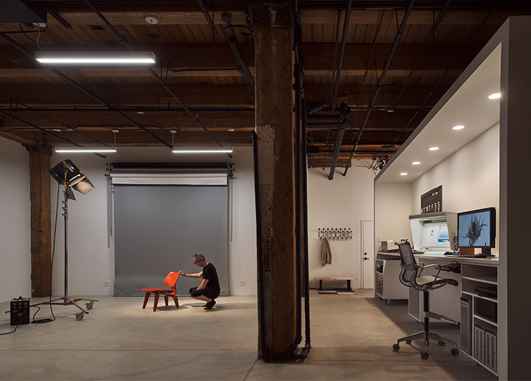 Christopher Barrett arranging a red chair under a spotlight in his Chicago studio, with timber beams and a workstation alongside