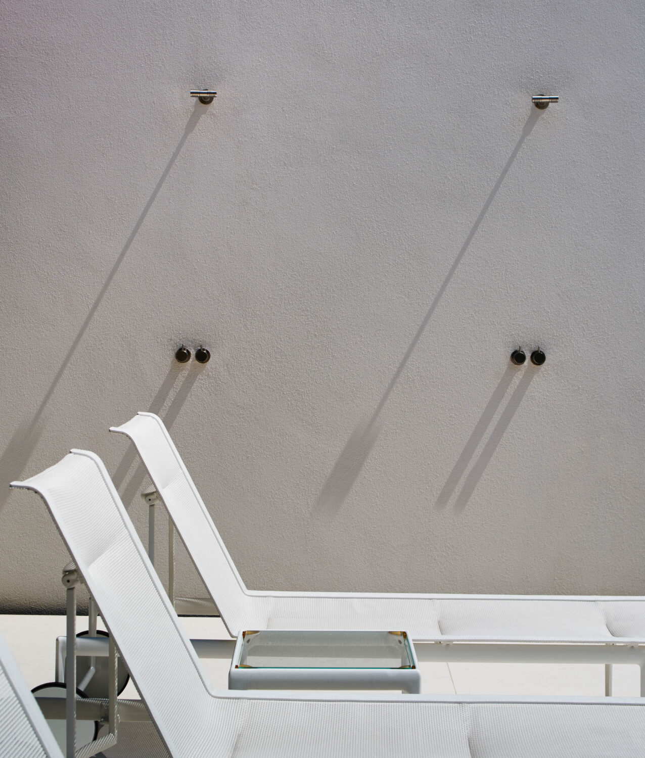 Outdoor shower fixtures casting long shadows on a white stucco wall above white chaise lounges