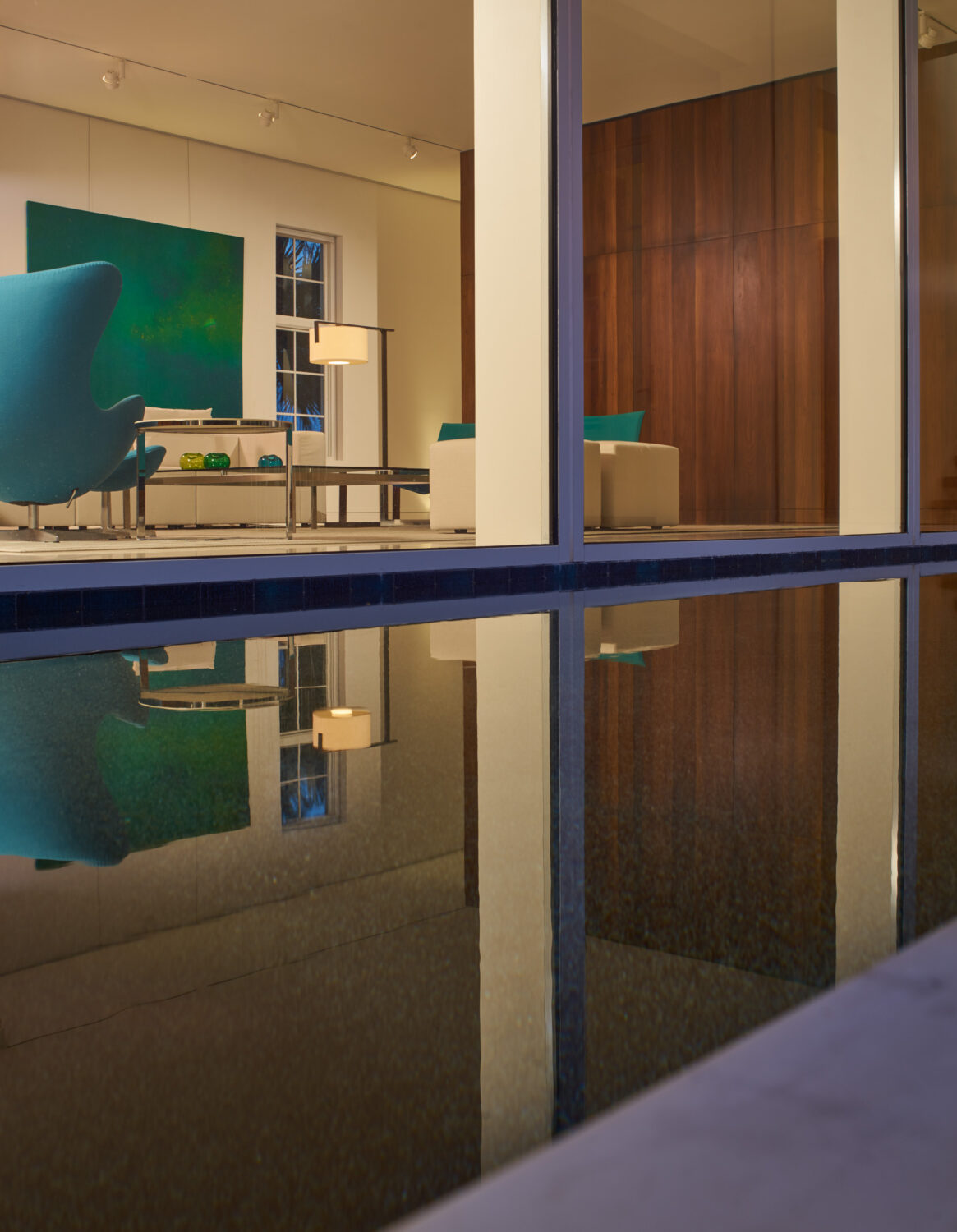 Living room seen through floor-to-ceiling glass and reflected in the courtyard pool at dusk