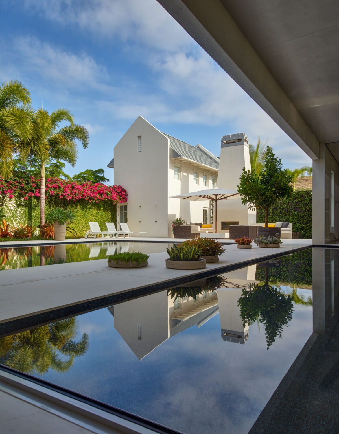 Guest wing, outdoor fireplace and palms reflected in the reflecting pool beneath a deep overhang