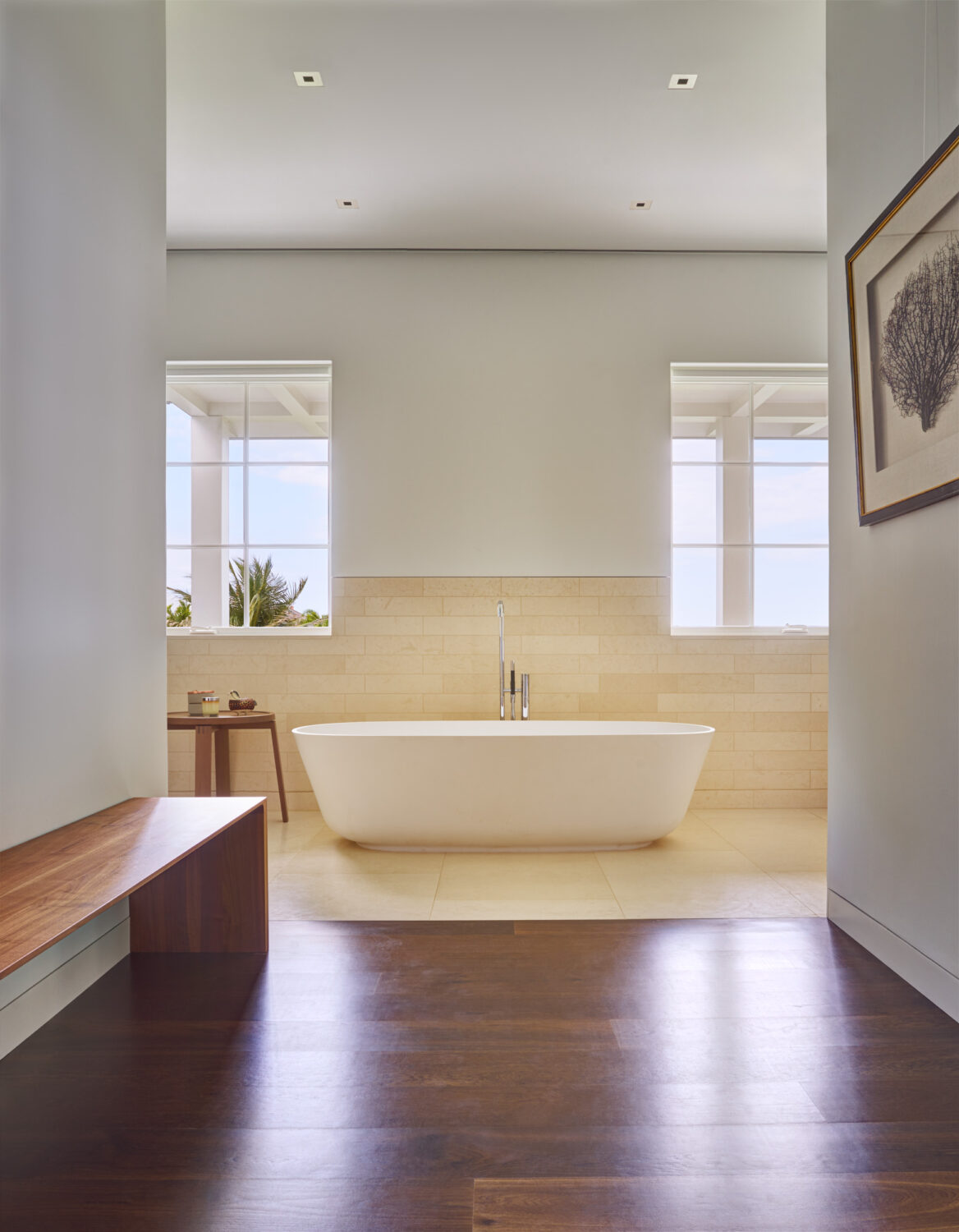 Freestanding white tub before a limestone wall between two windows in the primary bath