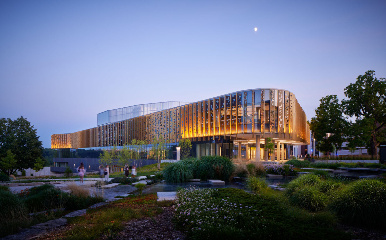 Western Michigan University Student Center at dusk, its gold perforated fins glowing above a landscaped garden with a pond and the moon overhead, Kalamazoo