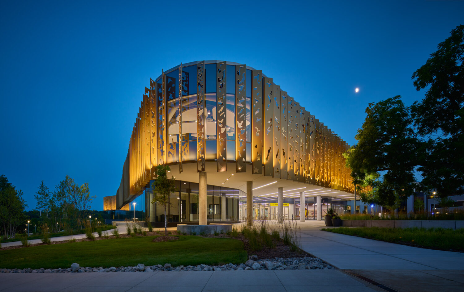 Curved corner of the Western Michigan University Student Center at blue hour, lit gold fins wrapping the glass above a column-supported entry