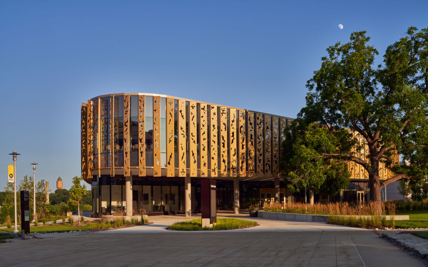 Rounded end of the Western Michigan University Student Center in early morning sun, gold cutout fins beside a large shade tree, Kalamazoo