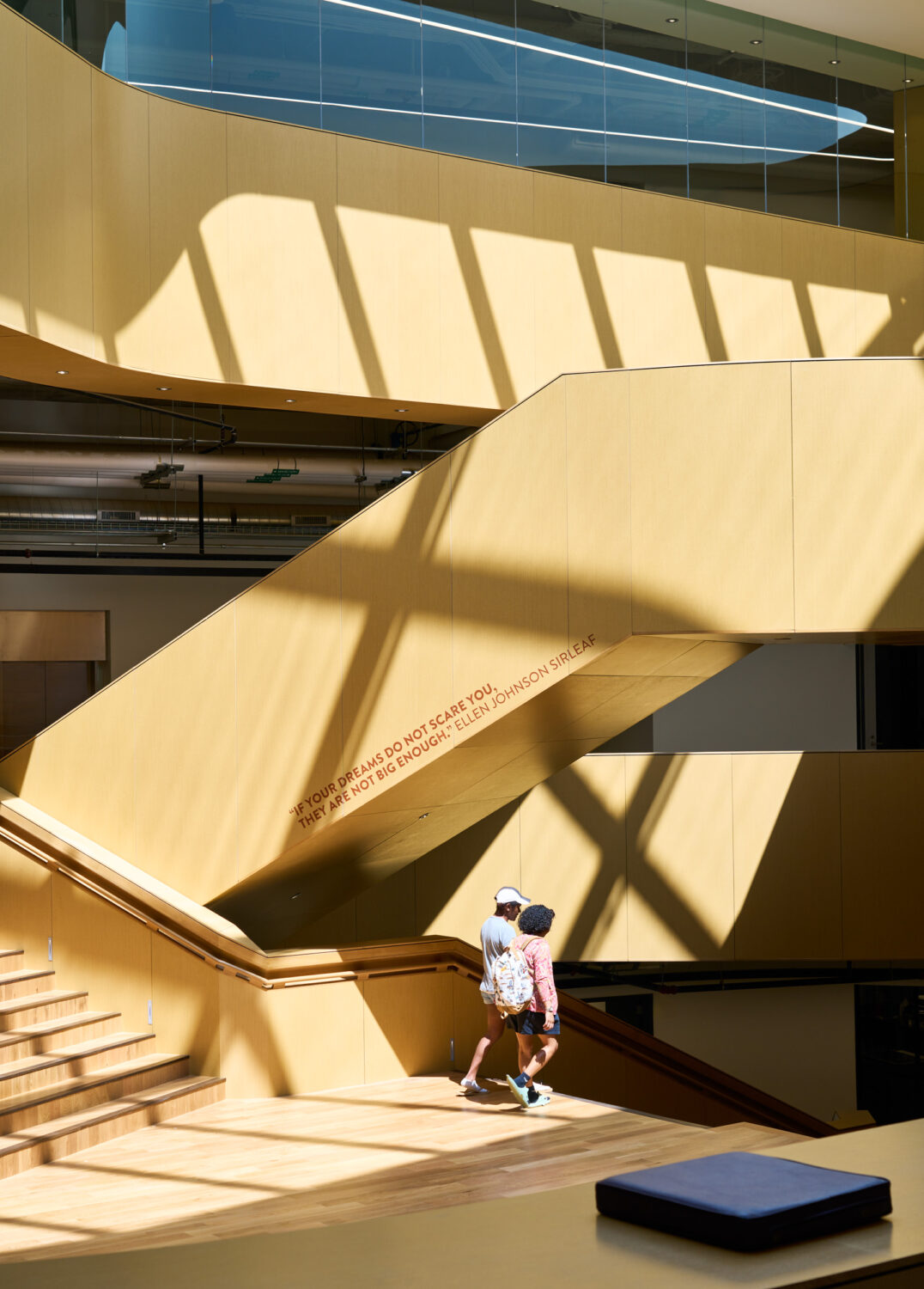 Sunlight casting diagonal shadows across the cedar stair walls of the Western Michigan University Student Center as two students climb