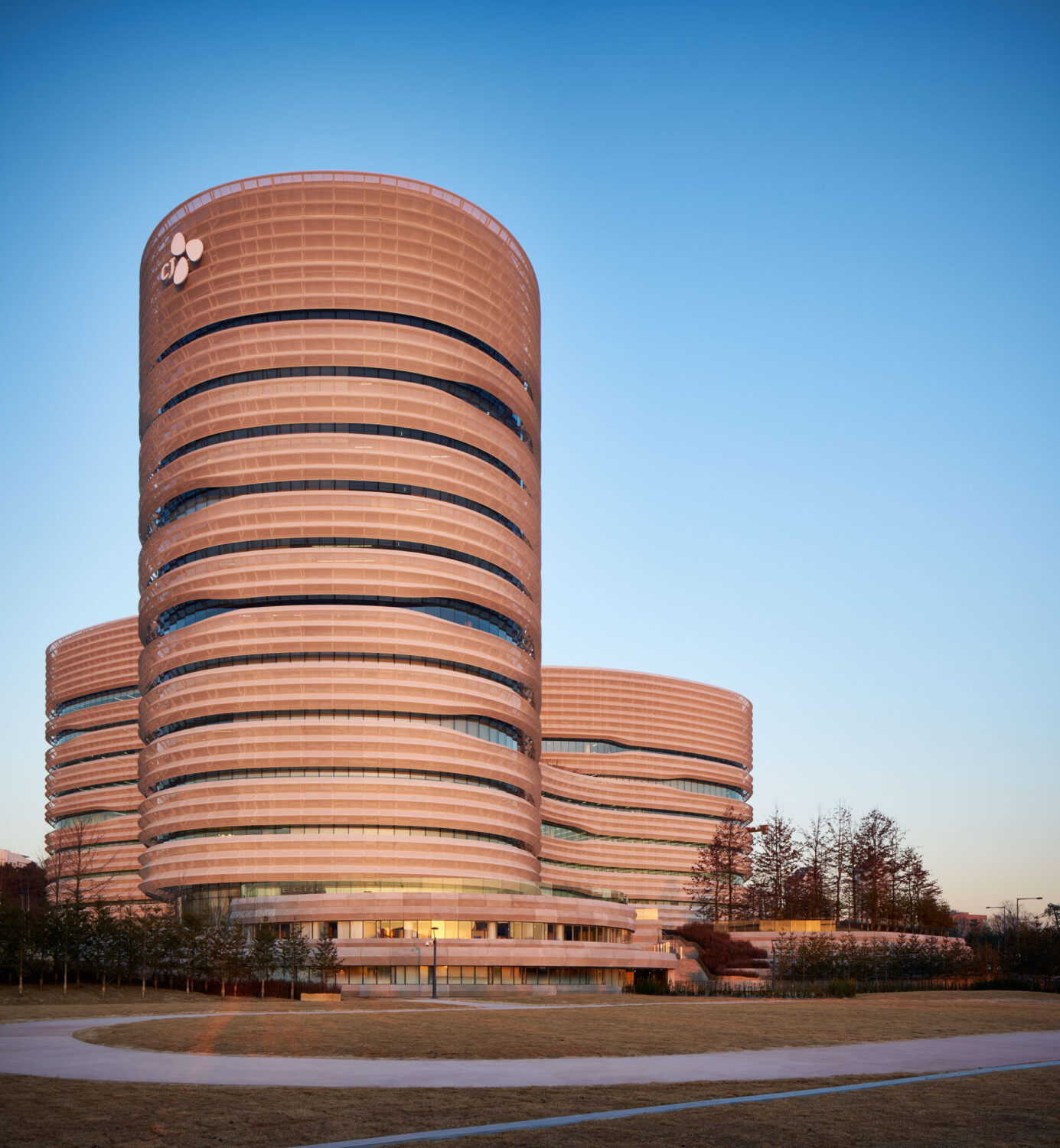 CJ Blossom Park's tallest elliptical tower at sunset, its perforated metal bands glowing against a clear blue sky, with two lower towers behind