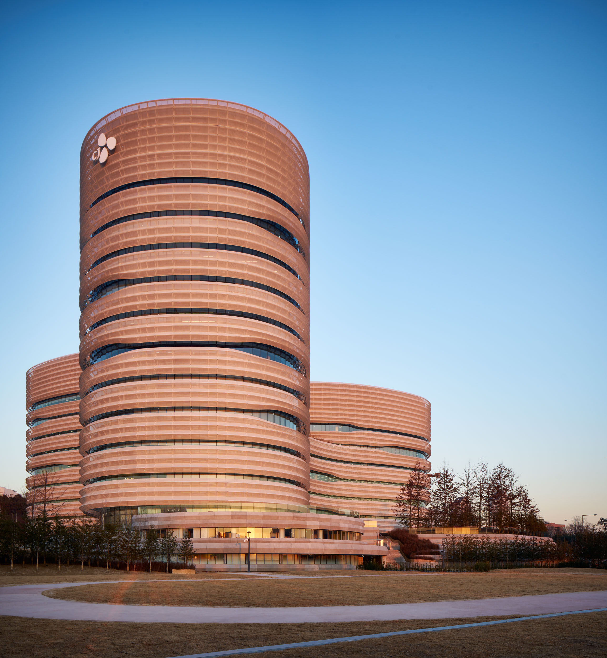 CJ Blossom Park's tallest elliptical tower at sunset, its perforated metal bands glowing against a clear blue sky, with two lower towers behind
