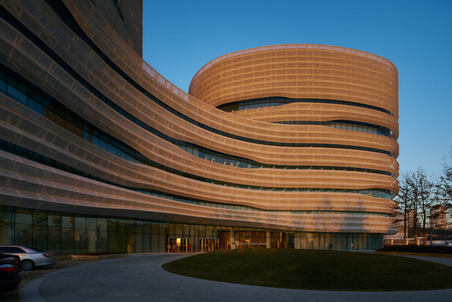 Curving perforated facade bands of CJ Blossom Park lit gold at sunset above the glass entrance and circular drive
