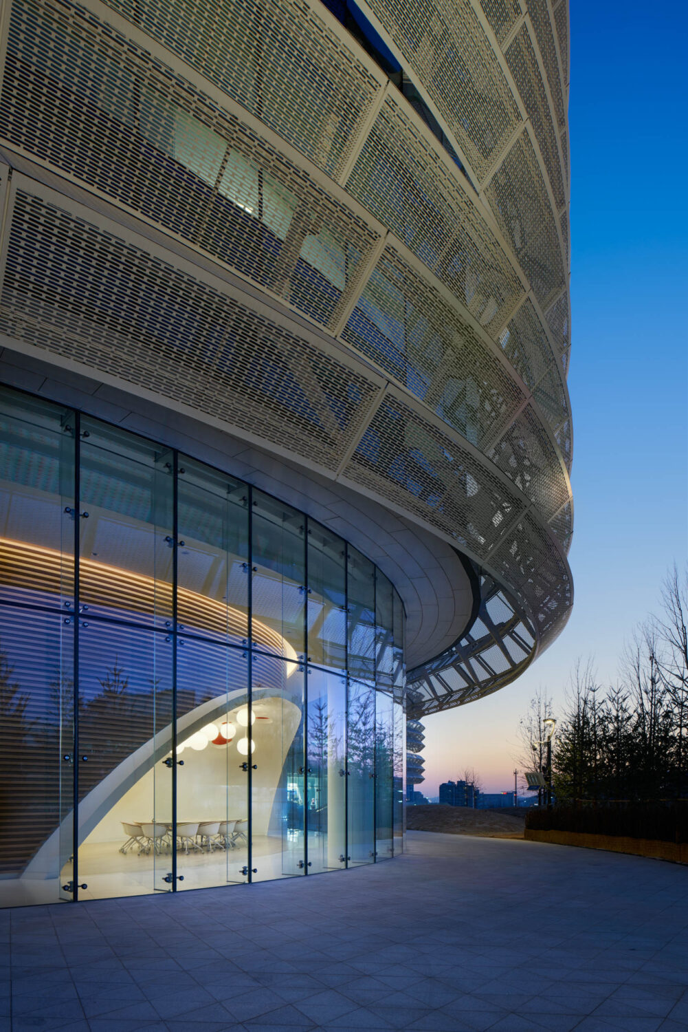 Perforated metal skin of CJ Blossom Park curving over a glass-walled meeting room at dusk