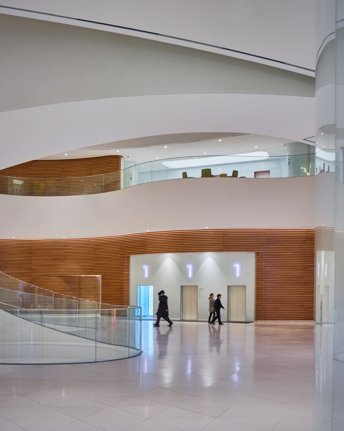 People walking past a wall of elevators framed in curved wood slats beneath white balconies at CJ Blossom Park
