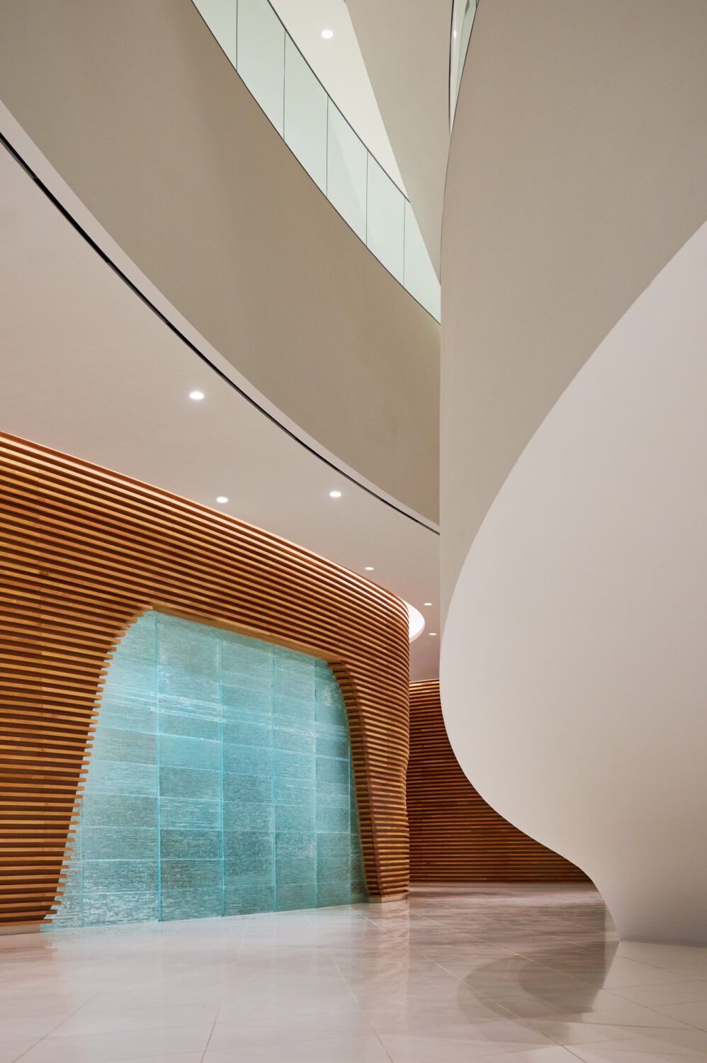 Cast glass water wall set into a curved wood slat wall beneath sweeping white balconies at CJ Blossom Park