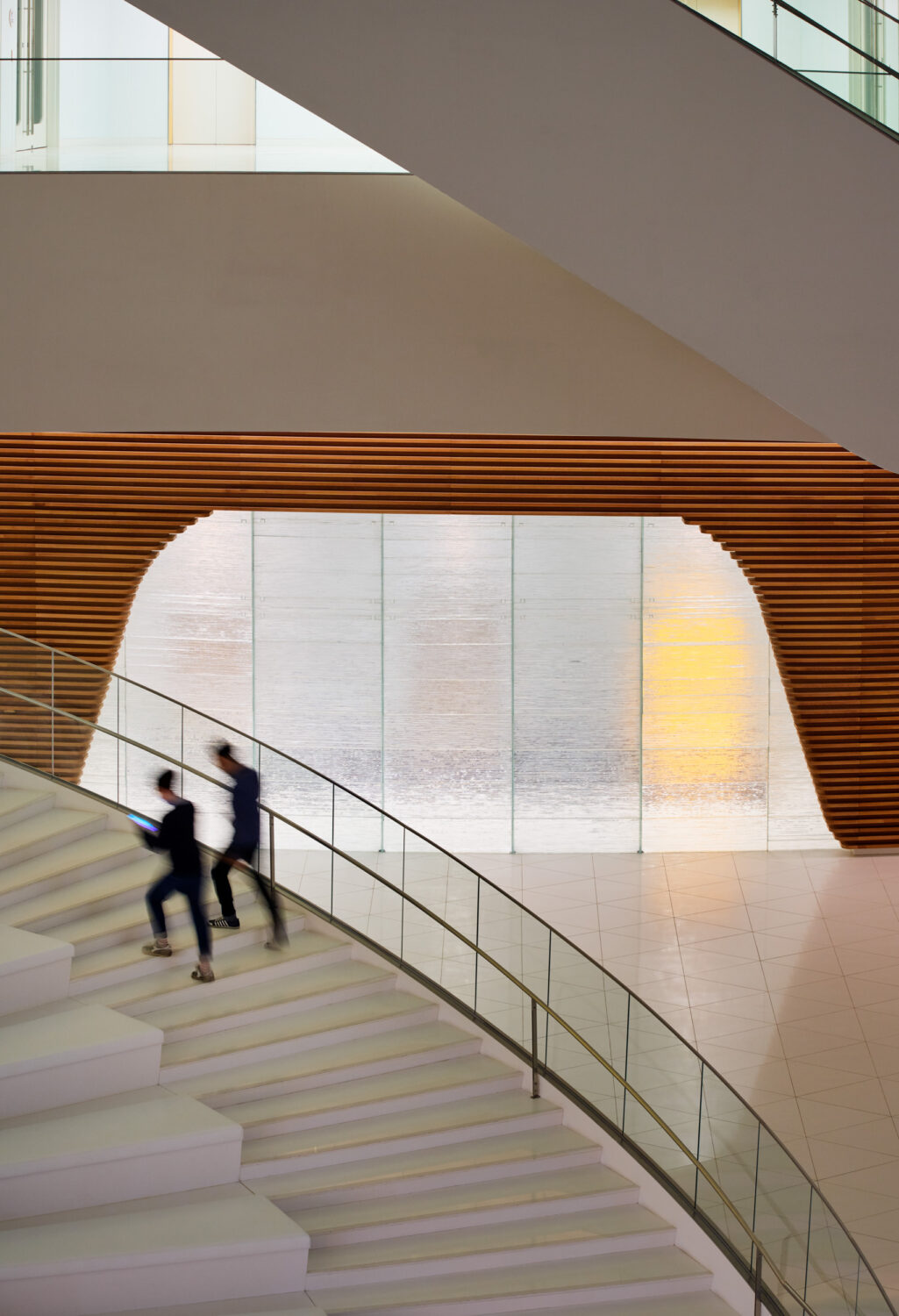 Two people climbing a white stair in front of a glowing glass wall framed in wood slats at CJ Blossom Park