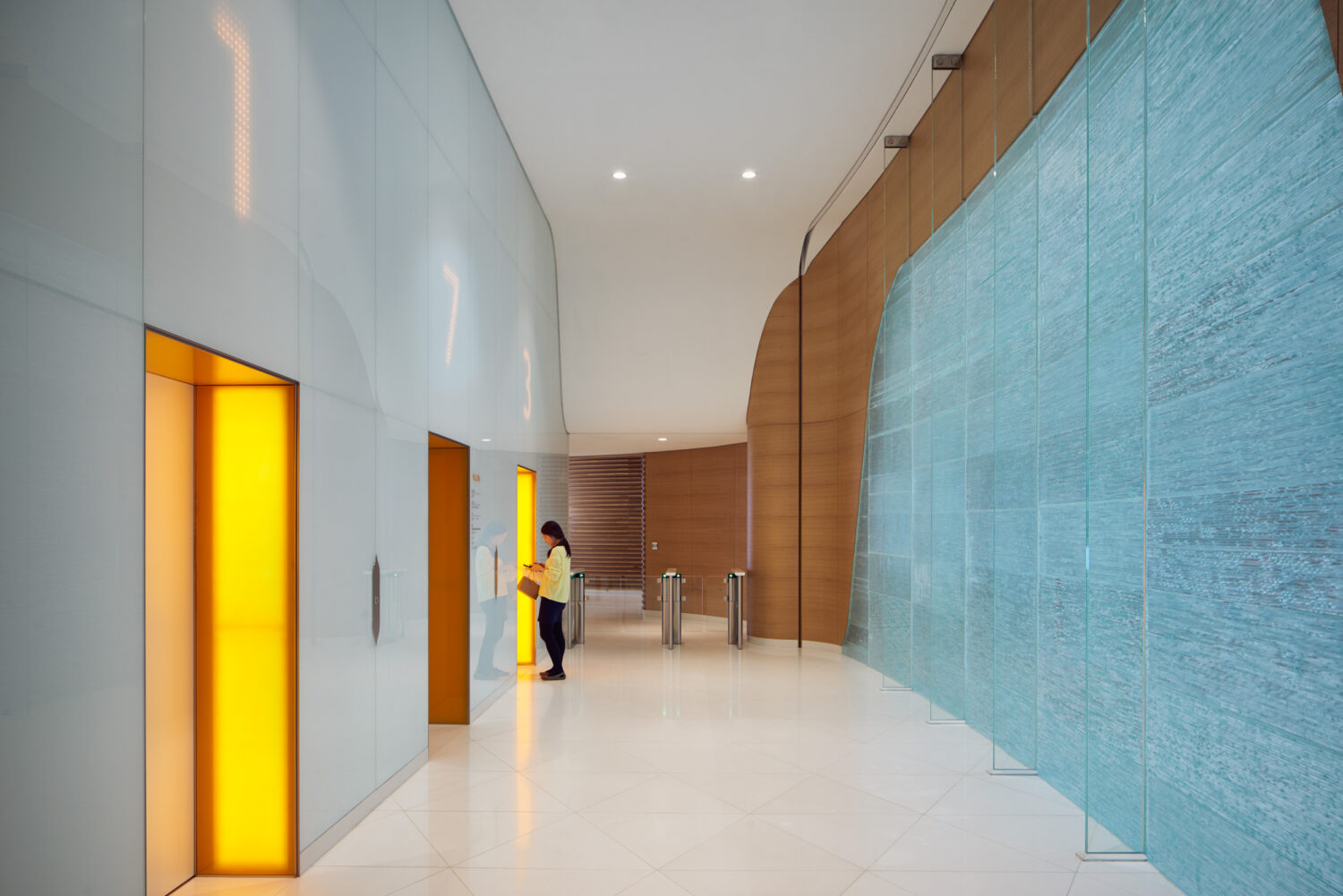 Elevator lobby at CJ Blossom Park with glowing yellow elevator doors opposite a wall of blue cast glass