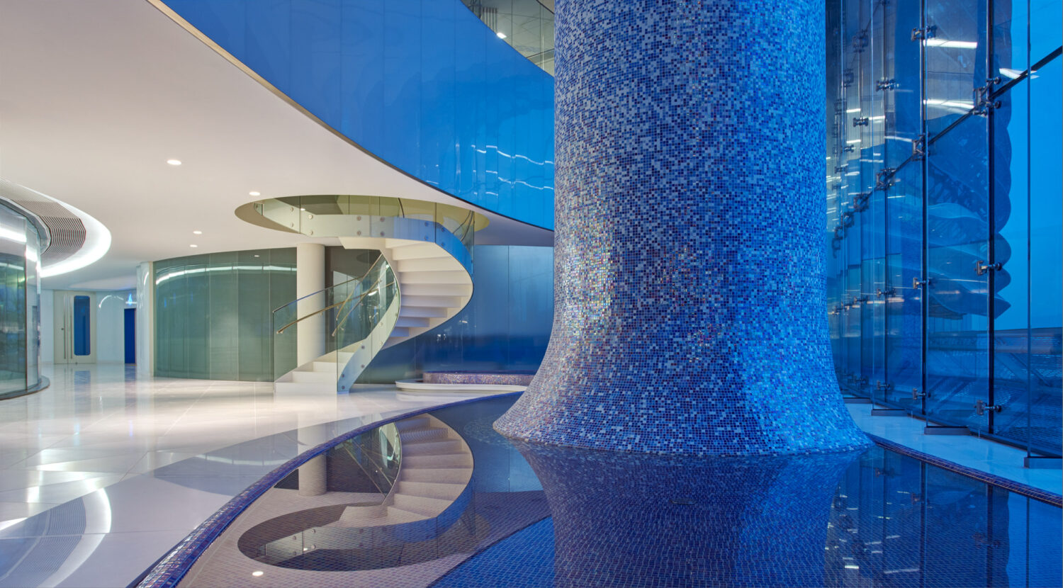 Blue mosaic-tiled column rising from a reflecting pool beside a spiral stair in the water garden at CJ Blossom Park