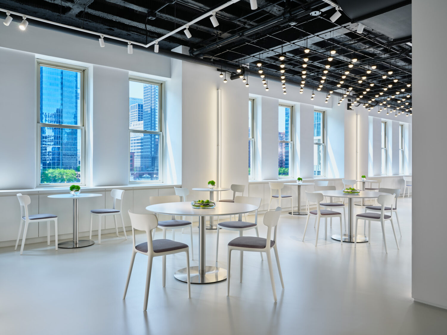 Rows of white cafe tables and chairs beneath a ceiling of pendant lights by tall windows in the Bernhardt showroom