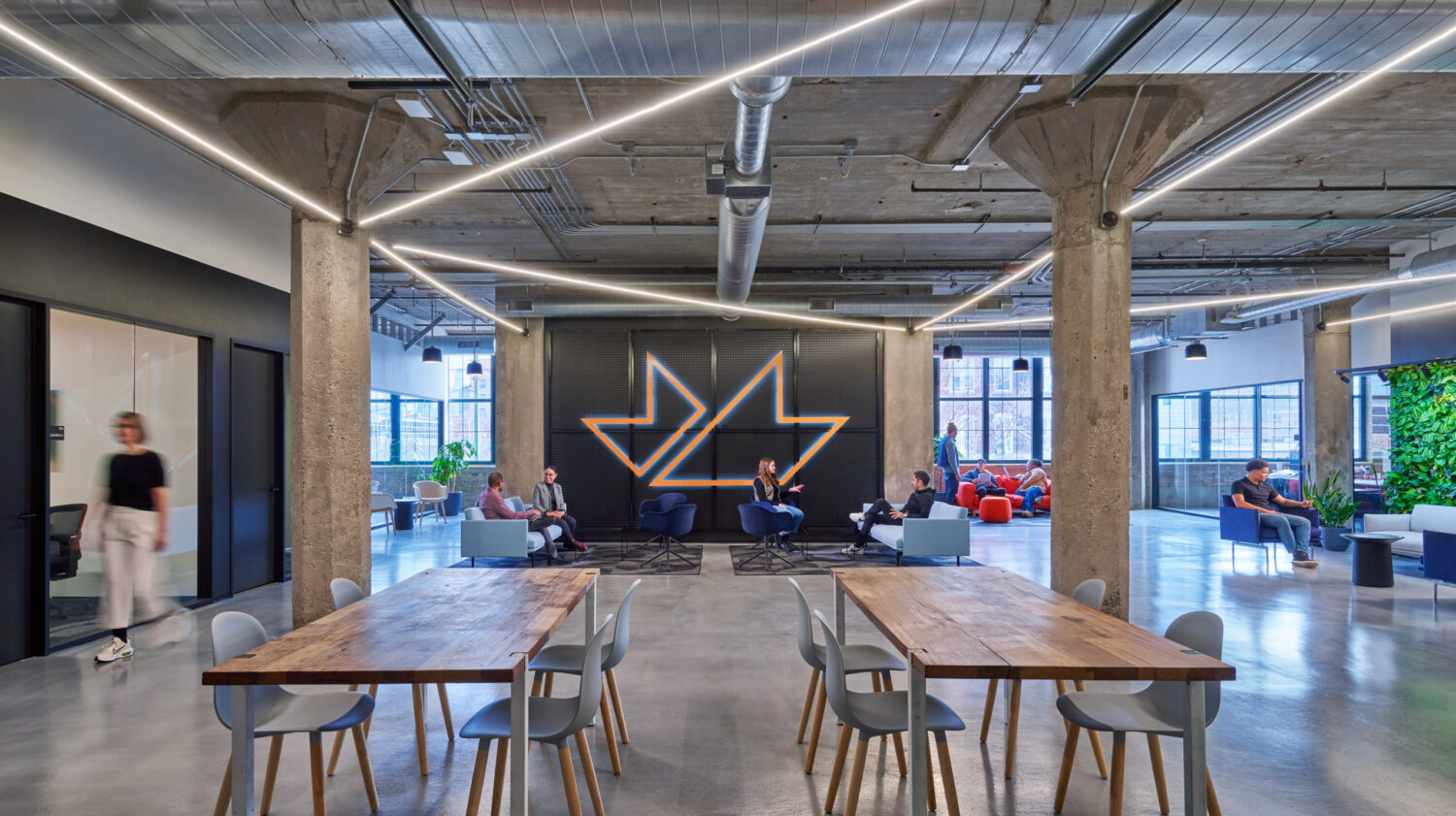Member lounge with long wood tables and concrete columns beneath the logo wall at mHUB