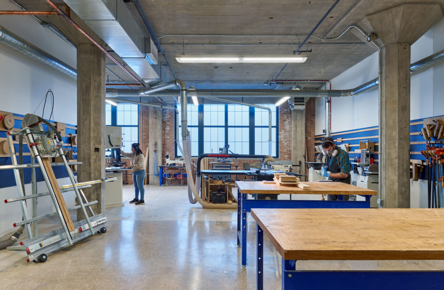 Woodworking shop with workbenches and a panel saw beneath tall factory windows at mHUB