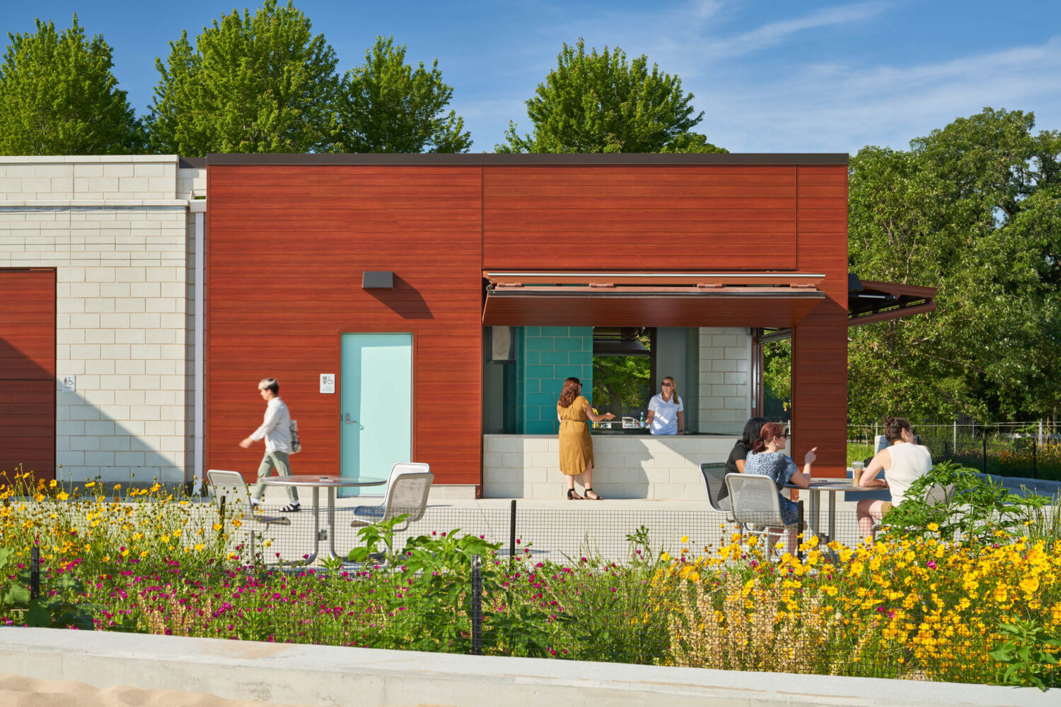 Visitors at the open concession window of South Shore Beach House, red wood-look panels and teal tile behind summer wildflowers