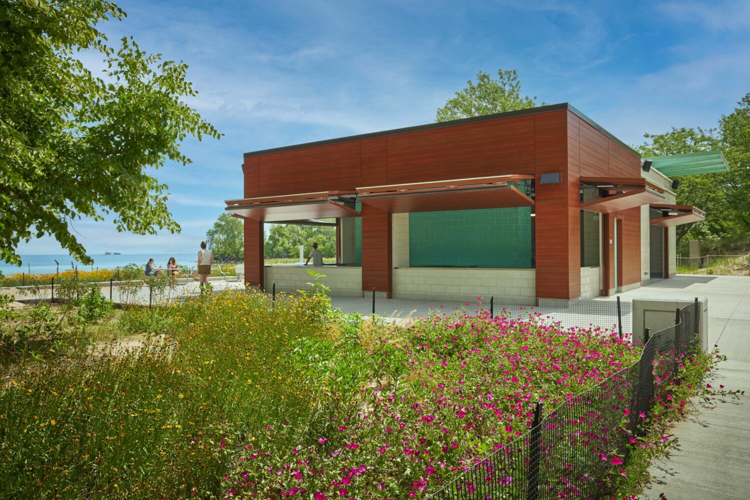 Corner of the South Shore Beach House concession pavilion with awnings raised toward Lake Michigan beyond pink wildflowers
