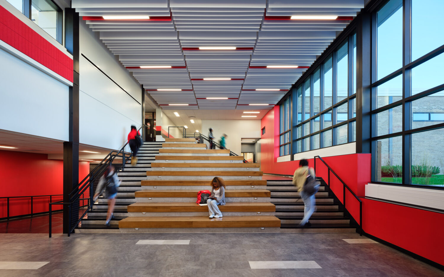Students sitting on wood bleacher steps beside a red stair in a double-height commons at Rich Township High School