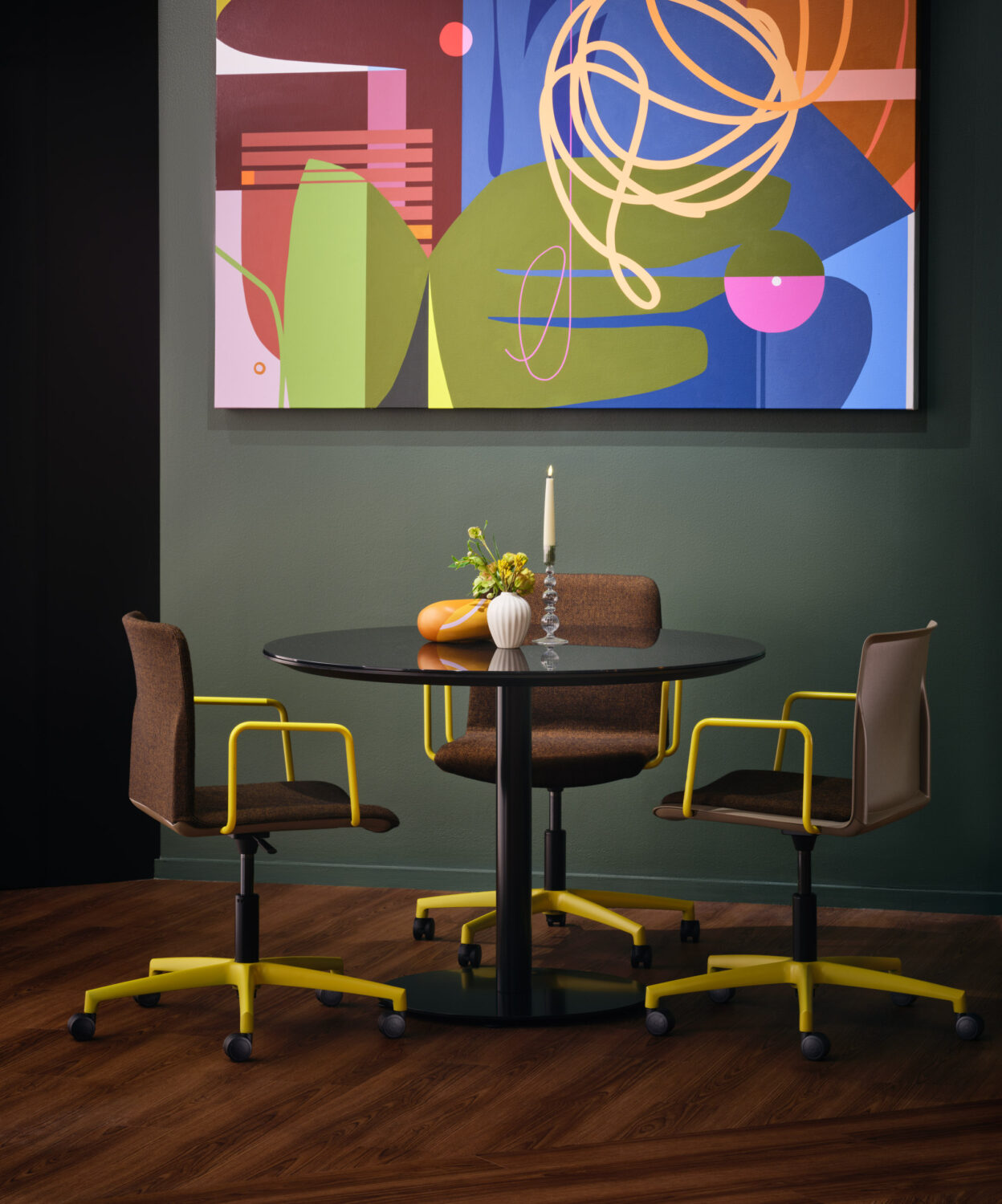 Small round table with dark chairs beneath a bright abstract painting on a deep green wall in the Source International showroom