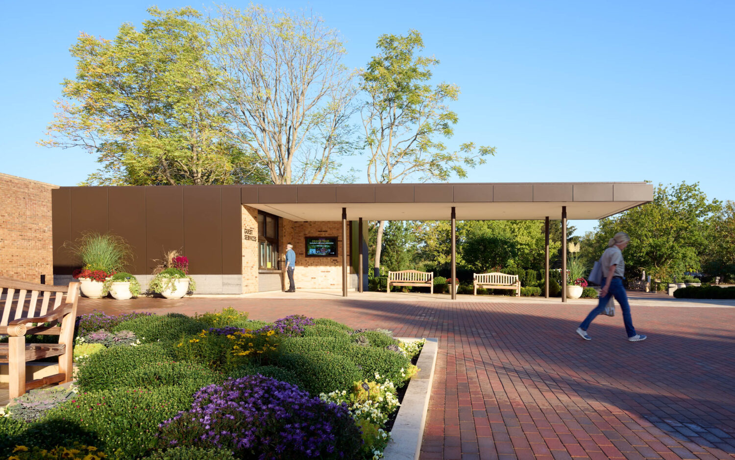 Long low canopy along the brick Welcome Plaza at the Chicago Botanic Garden, a visitor crossing beside the garden beds