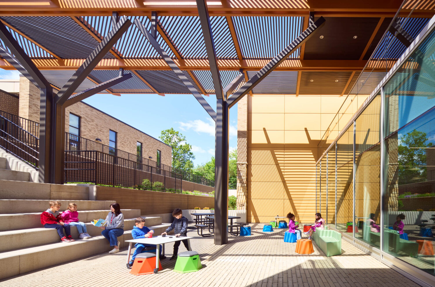 Children working at tables and on concrete steps beneath the slatted canopy of the outdoor amphitheater at the Kindergarten Center at Churchill Elementary