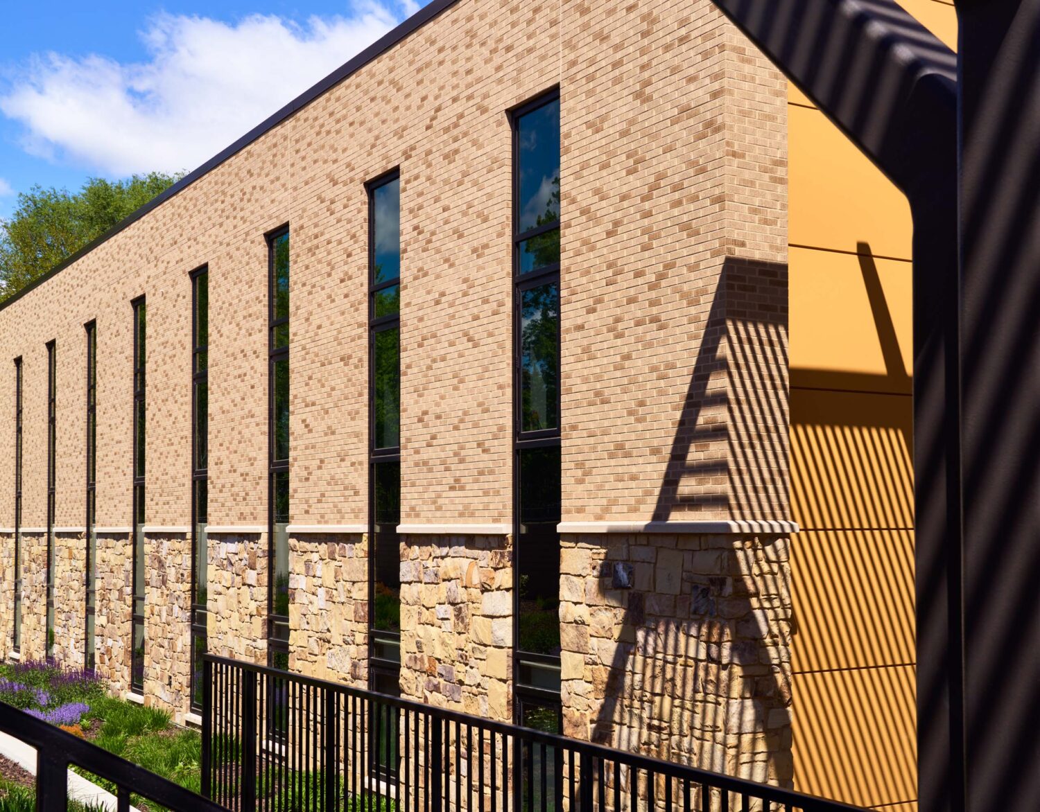 Brick and stone wall with tall narrow windows beside warm yellow panels striped with canopy shadows at the Kindergarten Center at Churchill Elementary