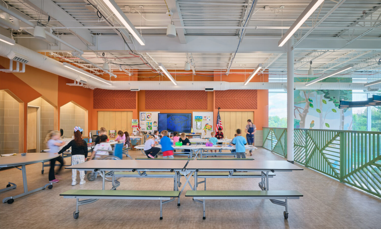 Upper-level dining commons with long tables, children at lunch and a green railing overlooking the multipurpose room at the Kindergarten Center at Churchill Elementary