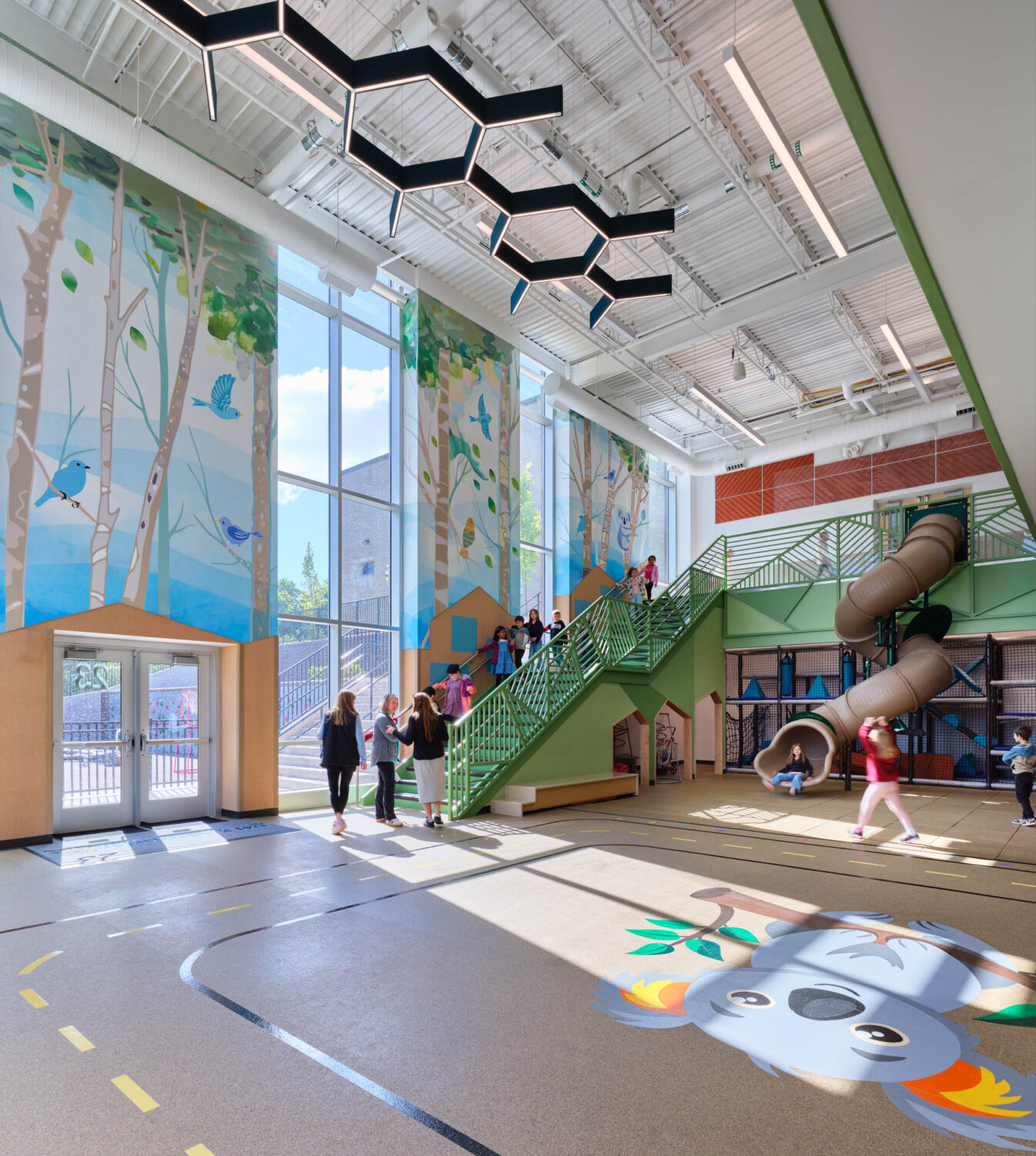 Two-story multipurpose room with tree murals, a green stair and a slide, and children playing on a koala floor graphic at the Kindergarten Center at Churchill Elementary