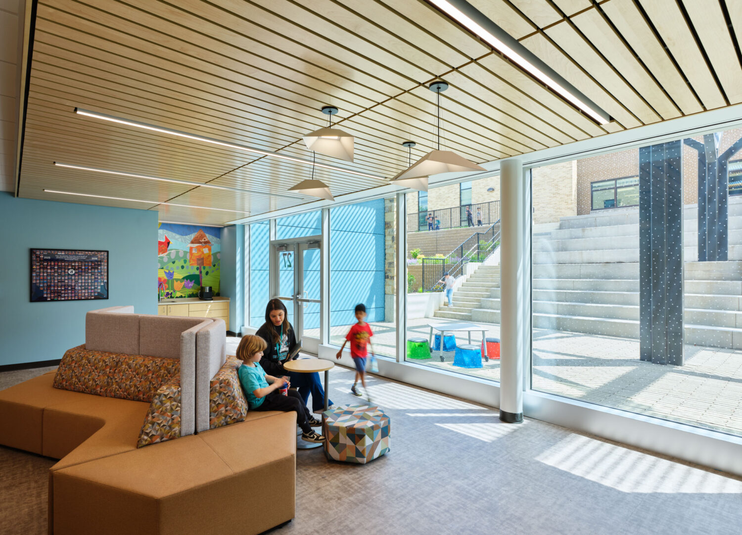 Sunlit lounge with curved seating and a teacher reading with children beside glass walls facing the courtyard steps at the Kindergarten Center at Churchill Elementary