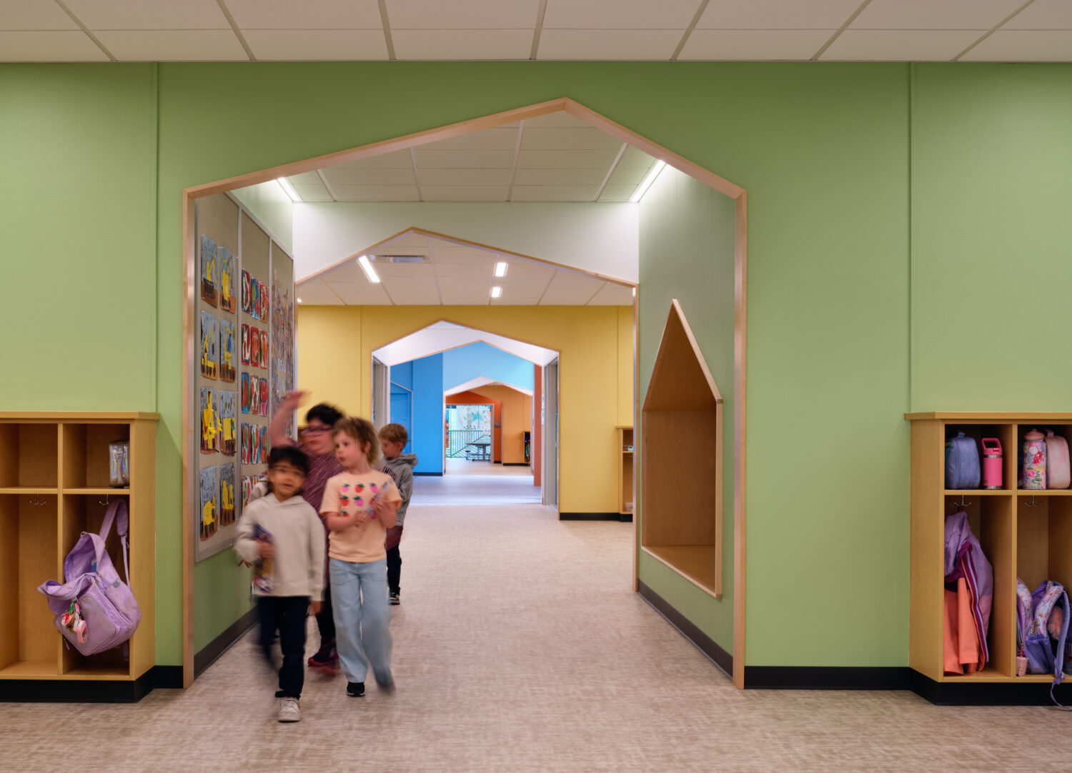 Children walking through a series of house-shaped portals in a green corridor lined with cubbies at the Kindergarten Center at Churchill Elementary