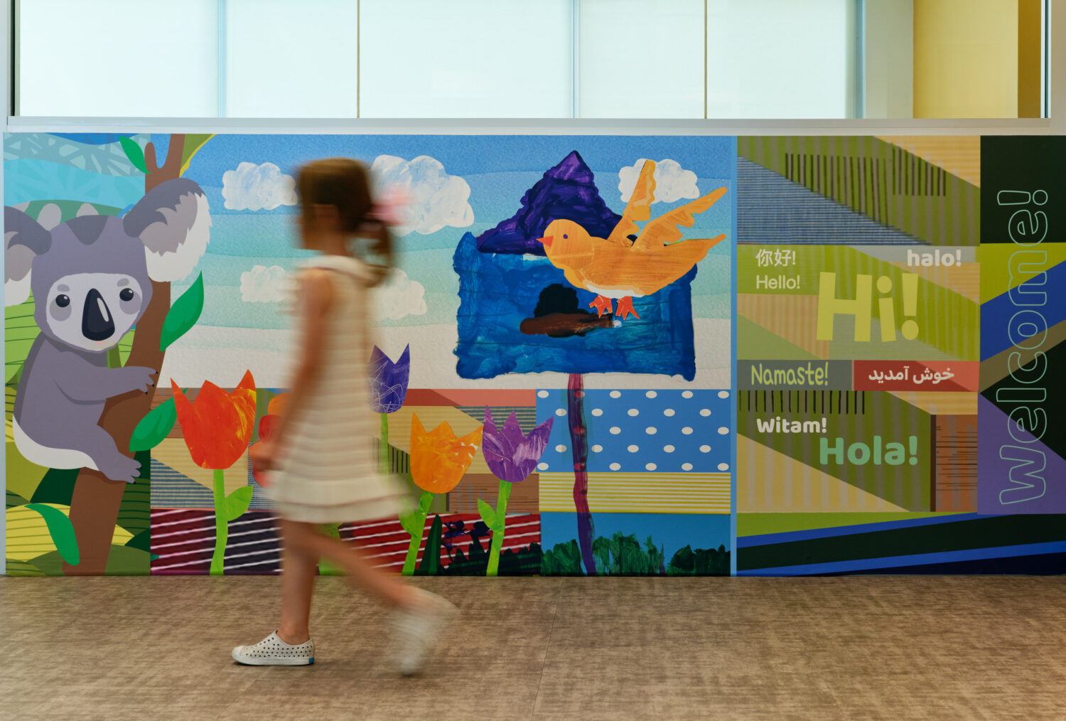 Girl walking past a colorful welcome mural with a koala and greetings in many languages at the Kindergarten Center at Churchill Elementary